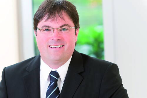 Lächelnder Geschäftsmann mit Brille im dunklen Anzug und gestreifter Krawatte vor einem hellen, unscharfen Bürohintergrund