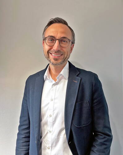 Lächelnder, mittelalter Mann mit Brille im dunklen Sakko und weißem Hemd steht vor einer hellen, neutralen Wand