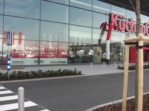 Moderne Glasfassade des Küchen Center Köln mit großen roten Schriftzügen, reflektierten Autos im Schaufenster und Fußgängerüberweg im Vordergrund