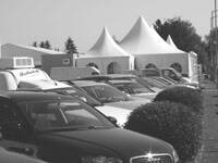 Reihe geparkter Autos entlang geschnittener Büsche vor großen weißen Eventzelten im Hintergrund