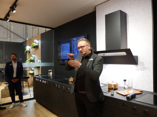 Mann mit Headset präsentiert in einer modernen Ausstellungs­küche eine schwarze Dunstabzugshaube und Kochfläche, während im Hintergrund ein Kollege und begrünte Wandmodule zu sehen sind.