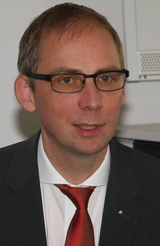 Mittelalterlicher Mann mit Brille im Anzug und roter Krawatte blickt lächelnd in die Kamera in einem Bürosetting