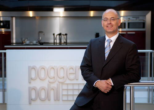 Geschäftsmann im Anzug lehnt lächelnd an einem Geländer vor einem modernen Küchenstudio mit großem „poggen pohl“-Firmenschild im Hintergrund
