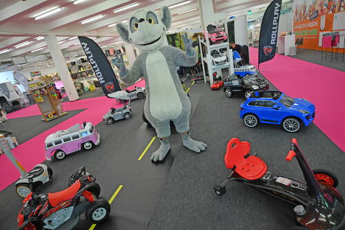 Große graue Maskottchenfigur begrüßt Besucher zwischen bunten Kinder-Elektroautos und Spielzeugfahrzeugen auf einer Messe mit pinkem Teppich.