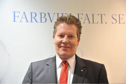 Geschäftsmann im Anzug mit orangefarbener Krawatte steht lächelnd vor einer hellen Wand mit dem teilweise sichtbaren Schriftzug „Farbvielfalt“
