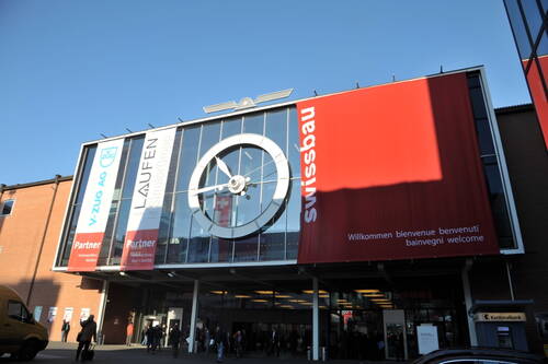 Eingangshalle eines Messegebäudes mit großer runder Uhr an der Glasfassade und roten Swissbau-Bannern, vor denen Besucher ein- und ausgehen.