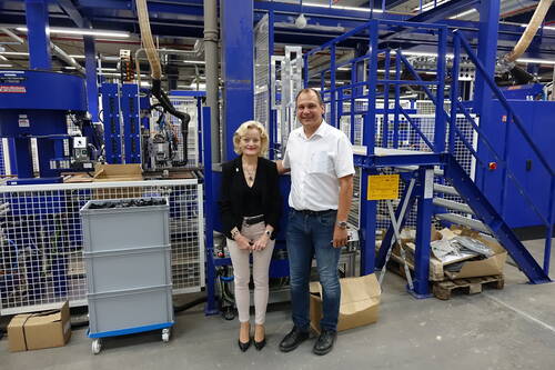 Zwei Beschäftigte stehen lächelnd in einer modernen Produktionshalle vor einer großen blauen Industrieanlage mit Treppe und Fördertechnik.