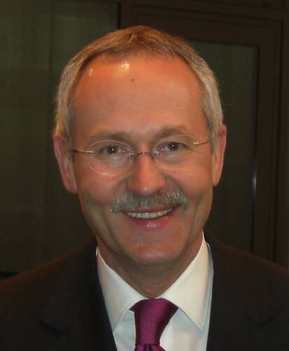 Älterer Mann mit Brille, grauem Schnurrbart und Anzug mit pinker Krawatte lächelt in die Kamera.