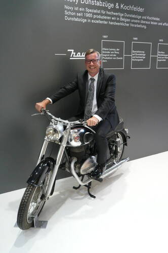Lächelnder Mann im Anzug sitzt auf einem klassischen silbernen Motorrad vor einer grauen Ausstellungswand mit Firmenhistorie und Text zu Dunstabzugssystemen.