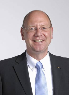 Älterer, lächelnder Mann mit Brille im dunklen Anzug und hellblauer Krawatte vor neutralem Hintergrund
