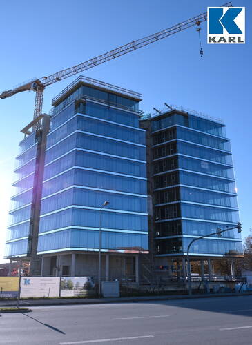Moderner Bürogebäudekomplex mit spiegelnder Glasfassade im Bau, daneben ein hoher Baukran vor blauem Himmel und Baustellenabsperrungen an der Straße.