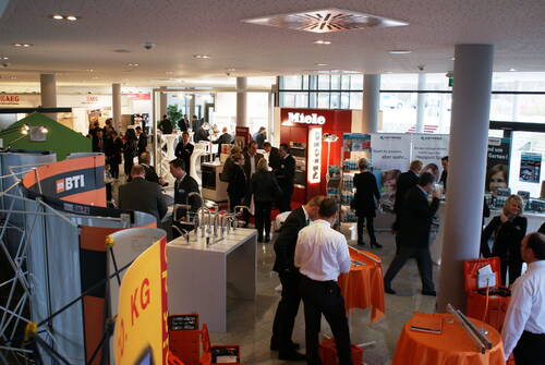 Besucher unterhalten sich und informieren sich an verschiedenen Ständen in einer hellen Messehalle mit Ausstellungen zu Haushalts- und Bauthemen.