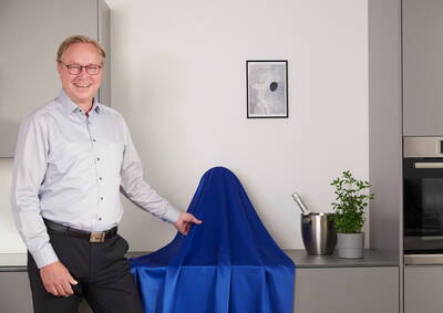 Lächelnder Mann in moderner Küche, der neben einem mit royalblauem Tuch verhüllten Gegenstand steht, flankiert von Champagnerkühler und Kräutertopf auf der Arbeitsplatte