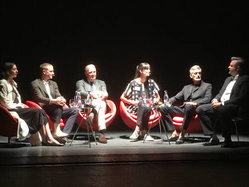 Sechs Personen sitzen in roten Sesseln auf einer dunkel beleuchteten Bühne und diskutieren konzentriert in einer Podiumsrunde.