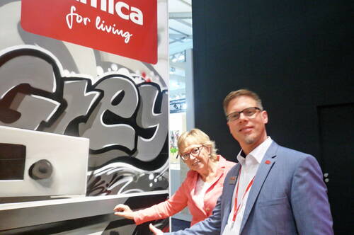 Zwei Messebesucher präsentieren lächelnd einen Küchenstand mit großem Graffiti-Schriftzug „Grey“ und einem roten Amica‑Werbeschild im Hintergrund.