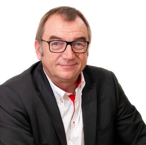 Mittelalterlicher Mann mit Brille in dunklem Anzug und weißem Hemd lächelt freundlich vor weißem Hintergrund