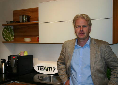 Mittelalter Mann im beigen Sakko lehnt an einer modernen Küchenzeile mit Kaffeemaschine, Spülbecken und TEAM7-Logo.