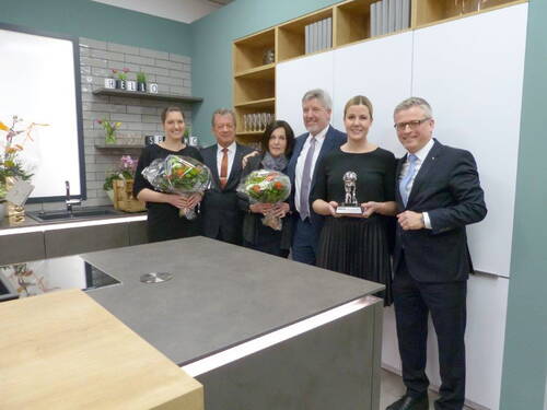 Sechs festlich gekleidete Personen stehen in einer modernen Einbauküche, zwei Frauen halten Blumensträuße und eine Frau präsentiert einen silbernen Preis-Pokal, während alle lächelnd in die Kamera blicken.