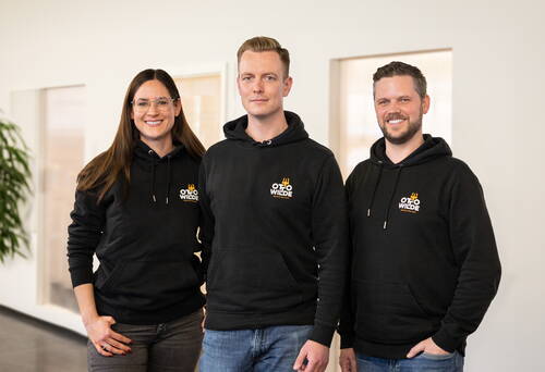 Drei lächelnde Teammitglieder in schwarzen „Otto Wilde“-Hoodies stehen nebeneinander in einem modernen Büro mit hellen Wänden und großen Fensterfronten.