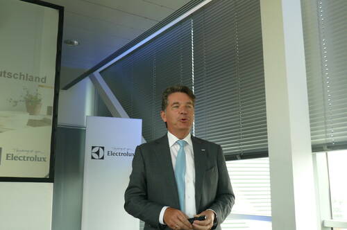 Geschäftsmann im grauen Anzug mit hellblauer Krawatte hält eine Präsentation vor Electrolux‑Werbetafeln in einem modernen Konferenzraum mit Jalousien.