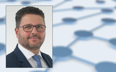 Mittelälterer Mann im Anzug mit Brille blickt freundlich in die Kamera, im Hintergrund ein unscharfes Netzwerkdiagramm