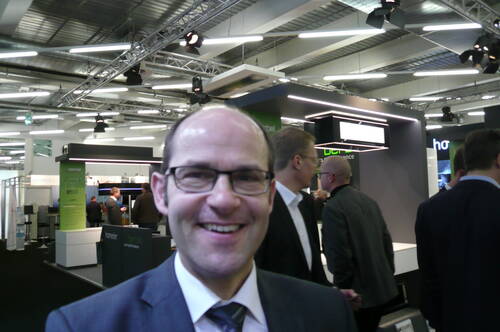 Lächelnder Mann mit Brille im Anzug steht im Vordergrund einer geschäftigen Messehalle mit mehreren Gesprächsgruppen und modernen Messeständen im Hintergrund