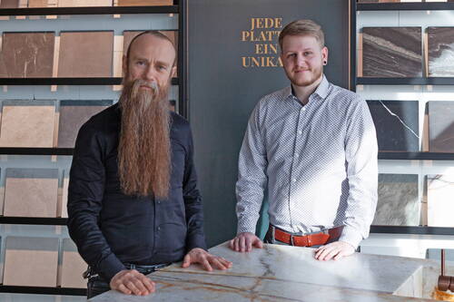 Zwei Männer stehen in einer Steinausstellung hinter einer polierten Marmorplatte, umgeben von Musterplatten verschiedener Natursteinoberflächen, im Hintergrund der Schriftzug „Jede Platte ein Unikat“.