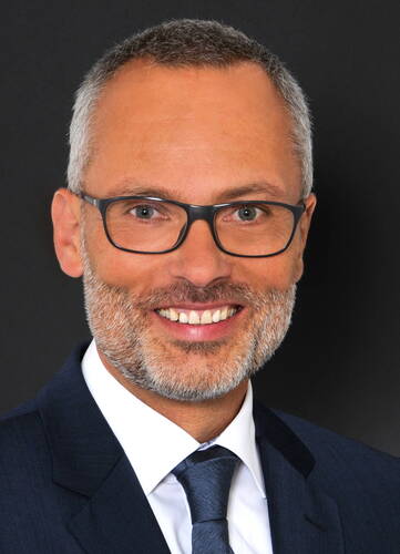 Lächelnder, mittelalter Mann mit graumeliertem Bart, Kurzhaarfrisur, dunkler Brille und dunkelblauem Anzug vor schwarzem Hintergrund