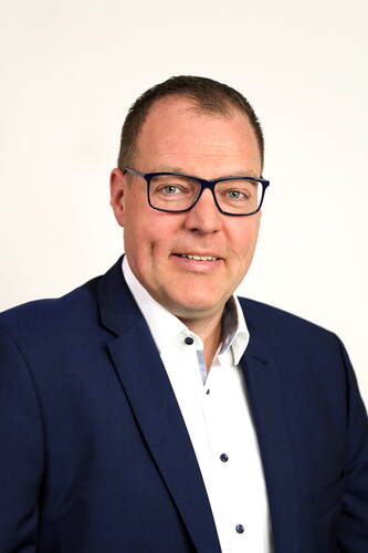 Mittelalterlicher Mann mit blauer Brille und dunkelblauem Anzug, der vor hellem Hintergrund freundlich in die Kamera lächelt