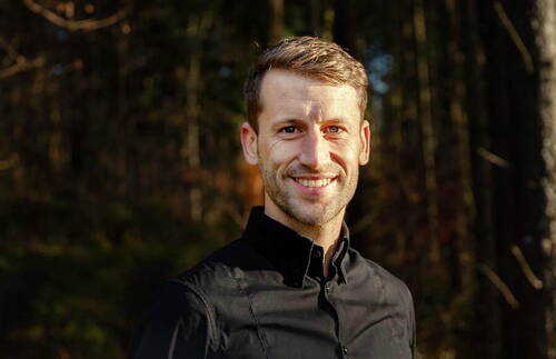 Junger Mann im schwarzen Hemd lächelt in warmem Sonnenlicht vor einem dunklen Waldhintergrund