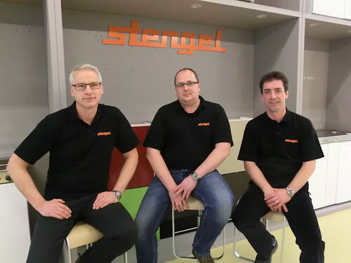 Drei Mitarbeiter in schwarzen Poloshirts mit orangefarbenem „stengel“-Logo sitzen in einer modernen Ausstellungsküche vor einem großen Firmen-Schriftzug.