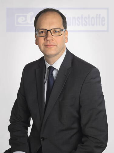 Geschäftsmann mit Brille im dunklen Anzug und Krawatte sitzt vor hellem Hintergrund mit unscharfem Firmenlogo.