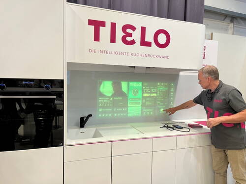 Mann demonstriert in einer modernen weißen Einbauküche eine interaktive, grün leuchtende Tielo-Küchenrückwand mit integrierter Touch-Bedienoberfläche.