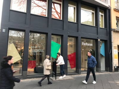 Passanten gehen an einem modernen Reform-Showroom mit großen Schaufenstern und bunten geometrischen Formen in einer städtischen Straße vorbei.