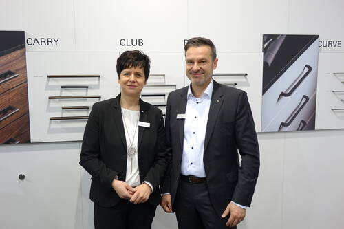 Zwei Geschäftsleute im Anzug präsentieren vor einer Messewand verschiedene moderne Griffleisten für Möbel und Schubladen.