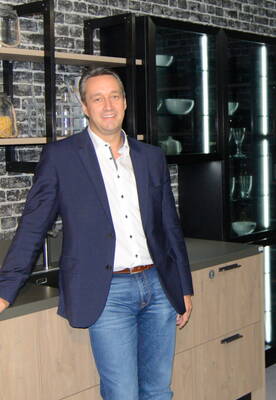 Mann im blauen Sakko und Jeans steht lächelnd in einer modernen Küche mit Backsteinwand, offenen Regalen und beleuchteten Glasschränken.