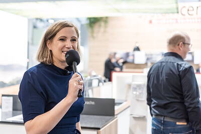 Lächelnde Moderatorin mit Mikrofon führt ein Interview in heller Messehalle mit Besuchern im Hintergrund