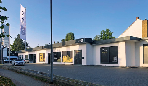 Moderner, eingeschossiger Showroom von cube30 mit großen Schaufenstern, Werbeaufschriften und Fahnen entlang eines leeren Parkplatzes an einer ruhigen Straße