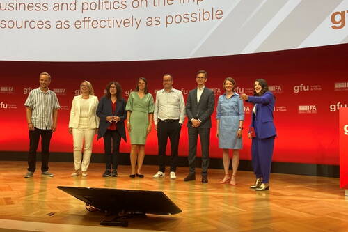 Neun lächelnde Konferenzteilnehmende stehen auf einer Bühne mit rotem Hintergrund und Präsentationsleinwand und posieren gemeinsam für ein Gruppenfoto.