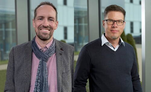 Zwei lächelnde Männer in Business‐Casual‐Kleidung stehen nebeneinander in einem modernen Bürogebäude mit Glasfassade im Hintergrund
