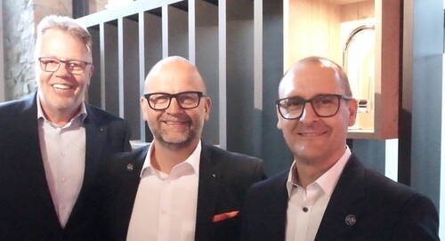 Drei lächelnde Männer im Anzug mit Brille stehen nebeneinander in einem modern beleuchteten Innenraum