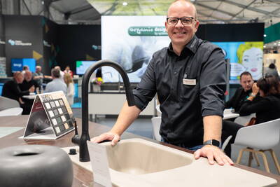Messeberater mit Namensschild präsentiert an einem Ausstellungsstand eine moderne Küchenspüle mit schwarzer Armatur, während im Hintergrund Besucher an Tischen Gespräche führen.