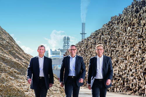 Drei Manager im Anzug gehen zwischen hohen Holz- und Hackschnitzelbergen in einem modernen Industrie- und Holzverarbeitungsbetrieb entlang
