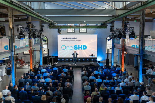 Redner präsentiert auf einer Bühne mit großem One-SHD-Display vor zahlreichem Publikum in einer industriell gestalteten Veranstaltungshalle