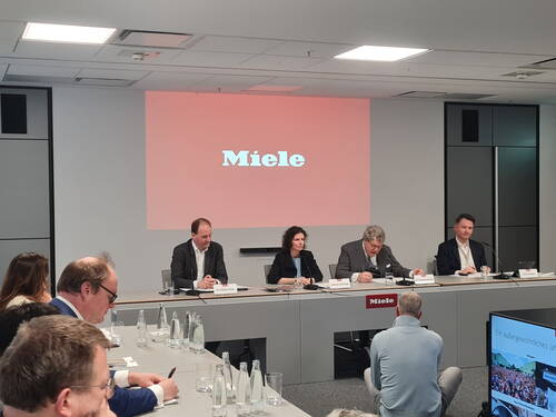 Managerpanel von Miele erläutert Geschäftszahlen in einem Konferenzraum vor sitzendem Publikum und großer Leinwand mit Unternehmenslogo im Hintergrund.