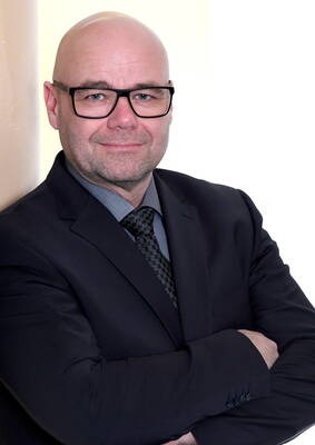 Glatzköpfiger Mann mit Brille im dunklen Anzug und gemusterter Krawatte steht mit verschränkten Armen vor hellem Hintergrund und blickt ruhig in die Kamera