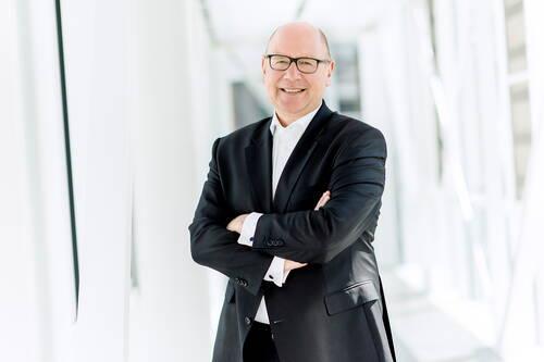 Geschäftsmann mittleren Alters im dunklen Anzug steht mit verschränkten Armen lächelnd in einem hellen, modernen Büroflur