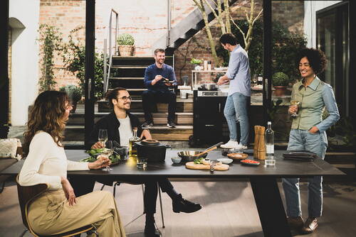 Freunde genießen gemeinsam ein entspanntes Abendessen in einem modernen Innenhof, während im Hintergrund am Grill gekocht und gelacht wird.