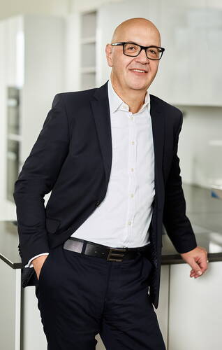 Glatzköpfiger Mann im dunklen Anzug und weißem Hemd lehnt entspannt an einer modernen hellen Büroküche und lächelt in die Kamera