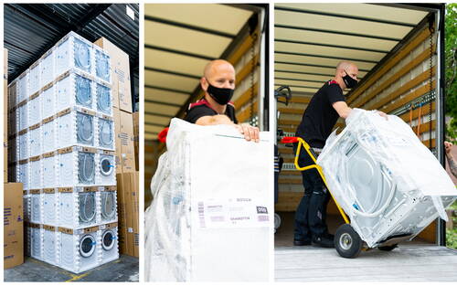 Mitarbeiter mit Mundschutz lädt verpackte Waschmaschinen auf einer Sackkarre aus einem Lkw, während im Lager hohe Stapel weiterer Geräte bereitstehen.